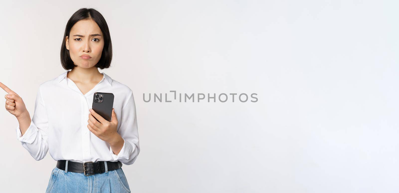 Skeptical asian modern girl, holding smartphone, pointing finger left at empty space and frowing hesitant, has doubts, standing over white background.