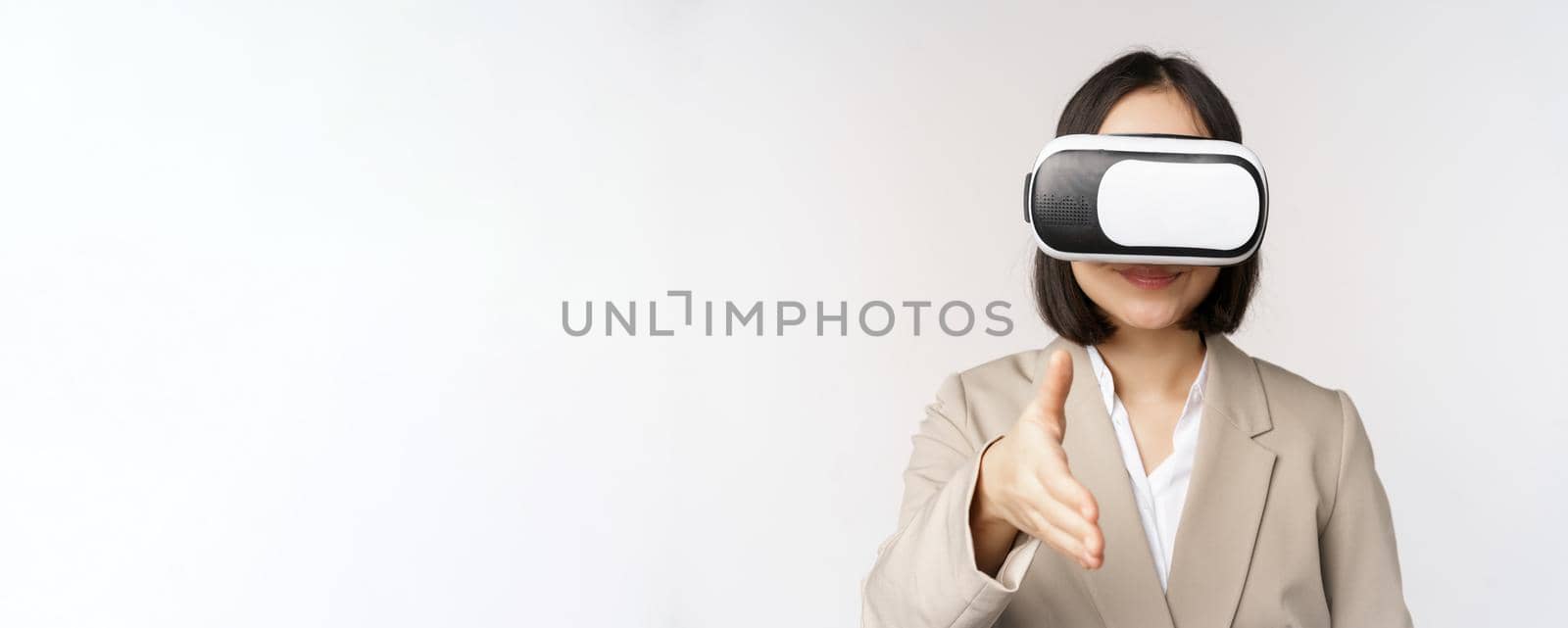 Meeting in vr chat. Asian businesswoman in virtual reality glasses, extending hand for handhshake with business partner, greeting someone, standing over white background by Benzoix