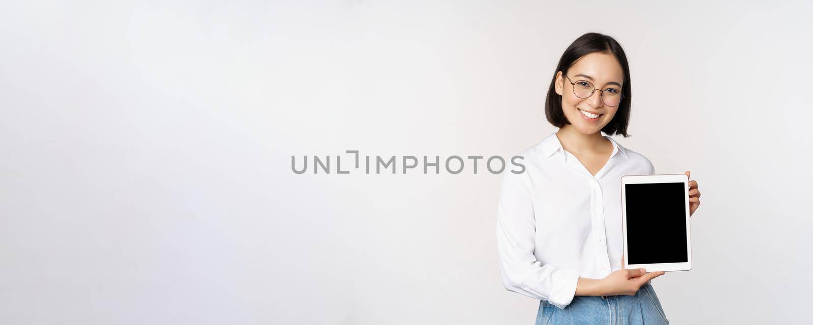 Young asian woman professional demonstrates digital tablet screen, info on her gadget, smiling and looking at camera, standing over white background by Benzoix