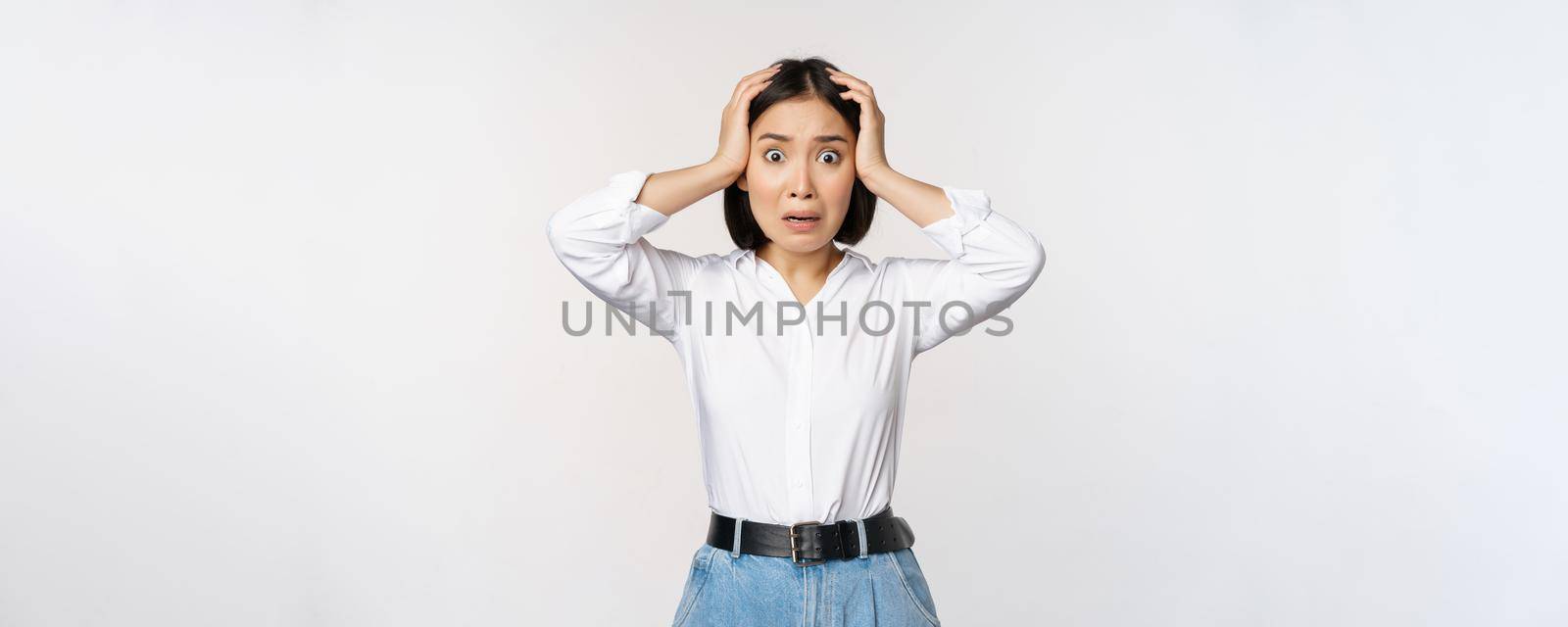 Image of shocked anxious asian woman in panic, holding hands on head and worrying, standing frustrated and scared against white background.
