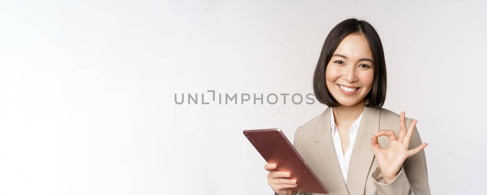 Image of successful asian businesswoman holding digital tablet, showing okay, ok sign, assuring client, standing over white background.