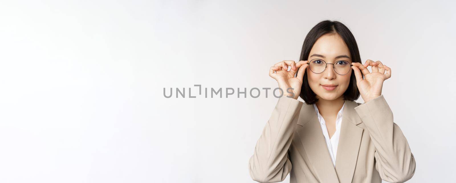 Smiling asian businesswoman trying new glasses, wearing eyewear, standing in suit over white background. Copy space