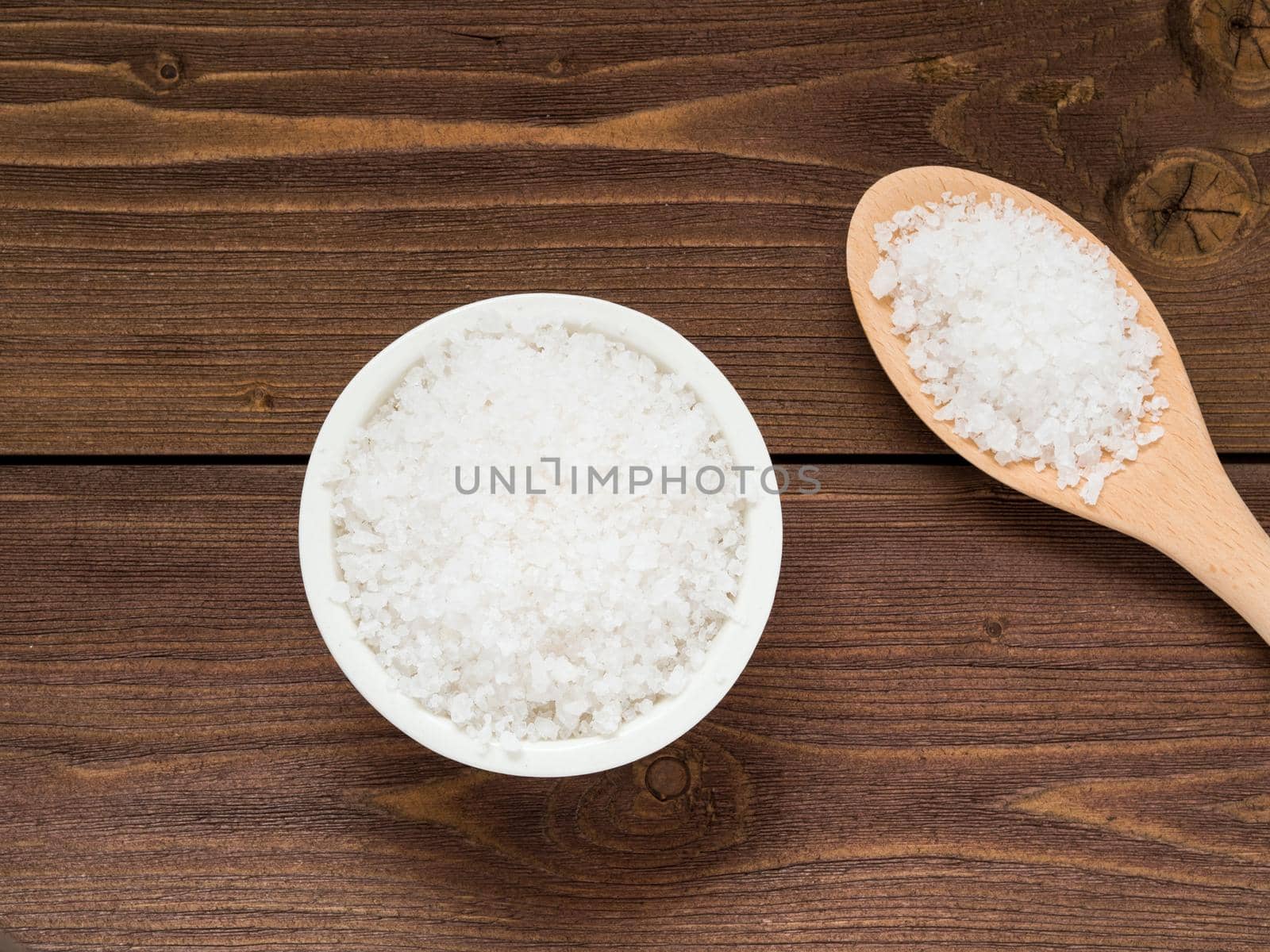 White bowl with large sea salt and wooden spoon on brown wooden table. Top view.