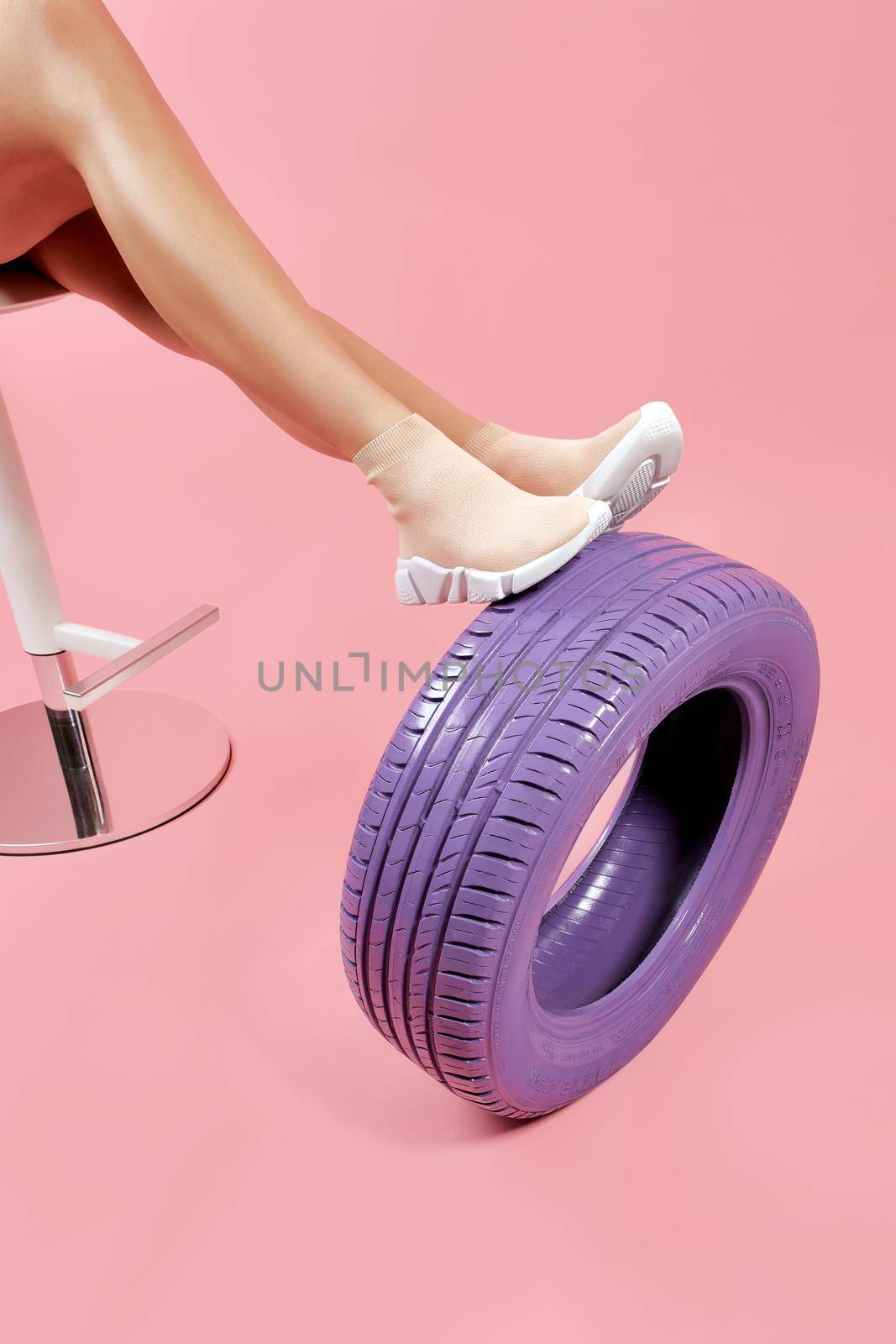 Slender female legs in elastic breathable beige sock sneakers with white soles leaning on light violet car tire on pastel pink background. Concept of trendy and comfortable sports style shoes