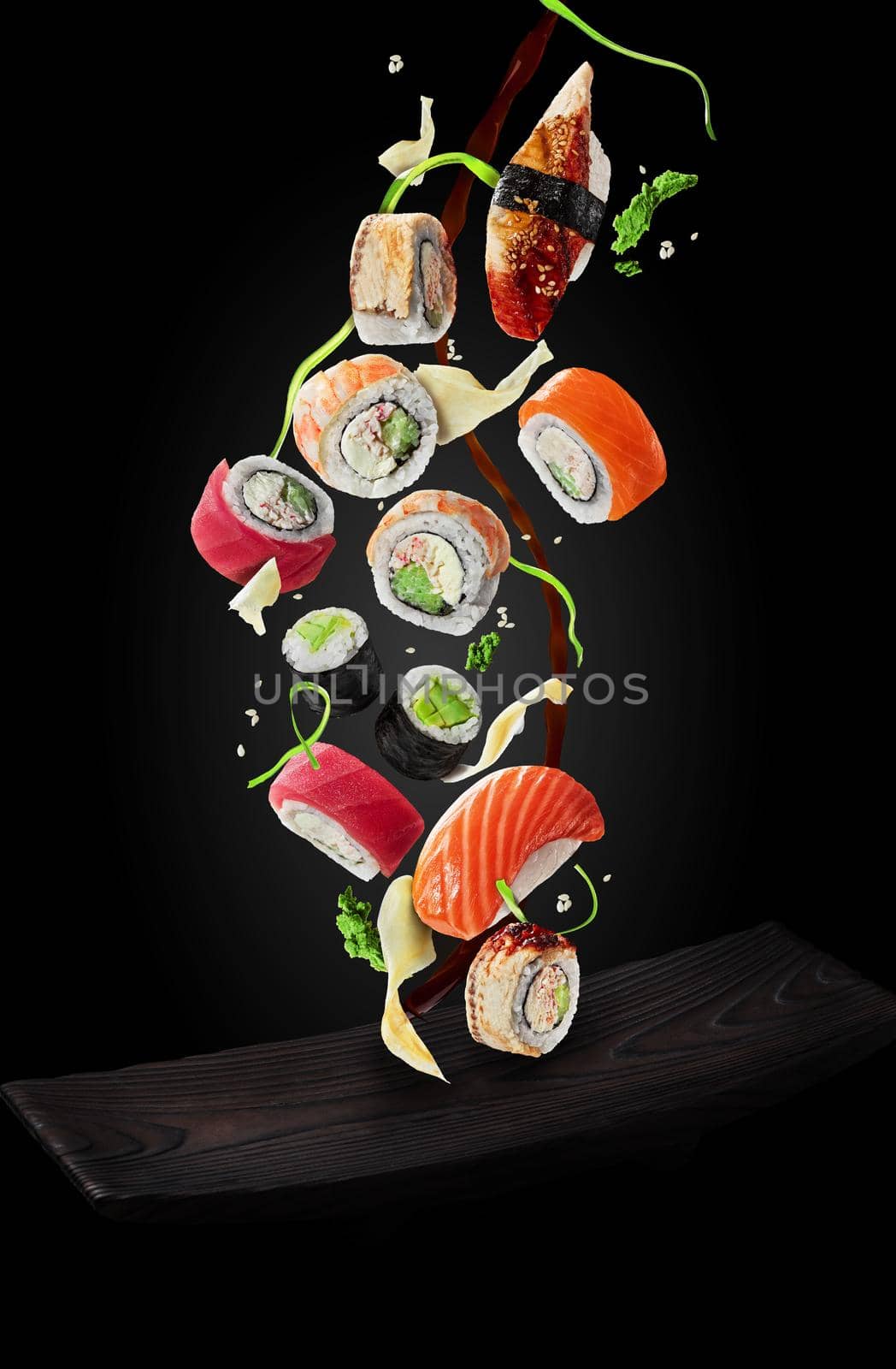 Set of various sushi with pieces of wasabi, slices of pickled ginger, sesame and jet of soy sauce hovering in air over wooden serving plate on black background. Food levitation concept