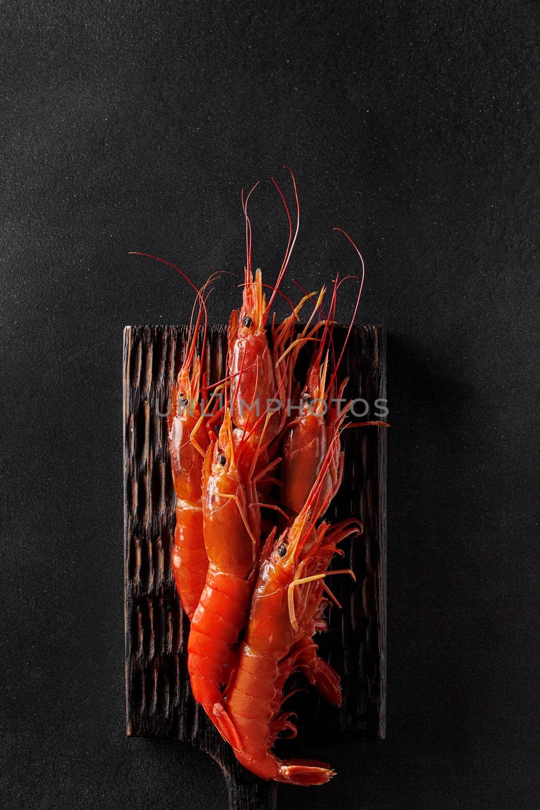 Top view of cooked whole red shrimps on wooden serving board on black background with copyspace. Vertical image