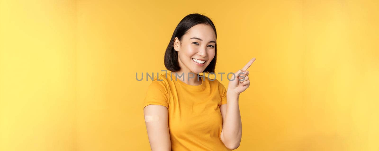 Vaccination and covid-19 pandemic concept. Smiling korean woman with band aid on shoulder after coronavirus vaccine shot, pointing at banner with vaccinating campaign by Benzoix