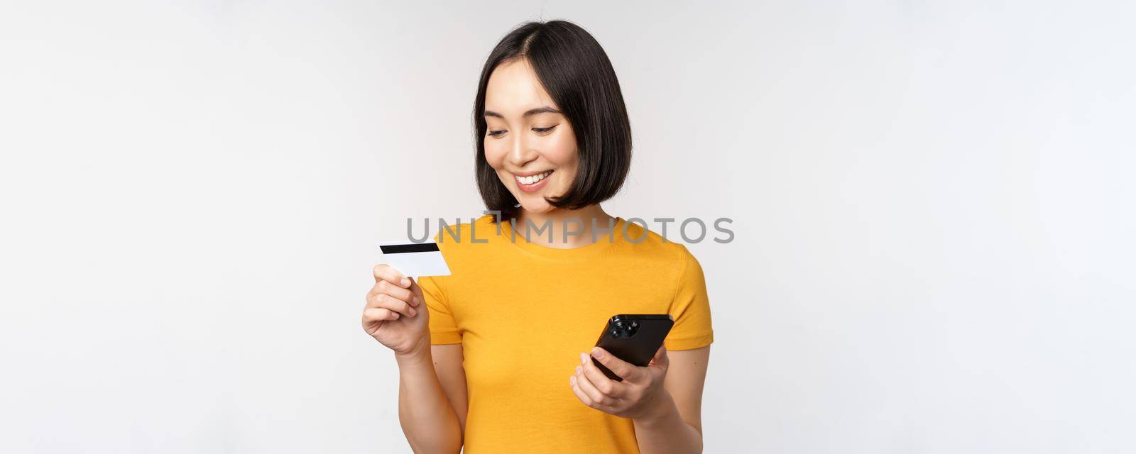 Beautiful smiling asian girl using credit card and mobile phone, paying online on smartphone, standing in yellow tshirt over white background by Benzoix