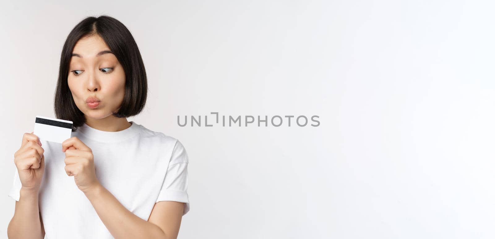 Money and finance concept. Cute japanese girl kissing her credit card, standing in tshirt over white background.
