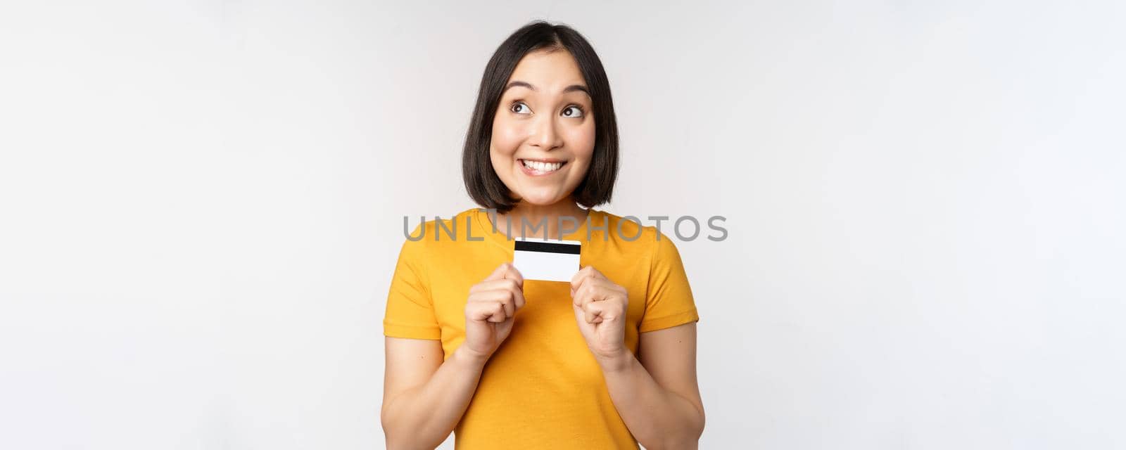 Portrait of beautiful korean girl holding credit card, recommending bank service, standing in yellow tshirt over white background.