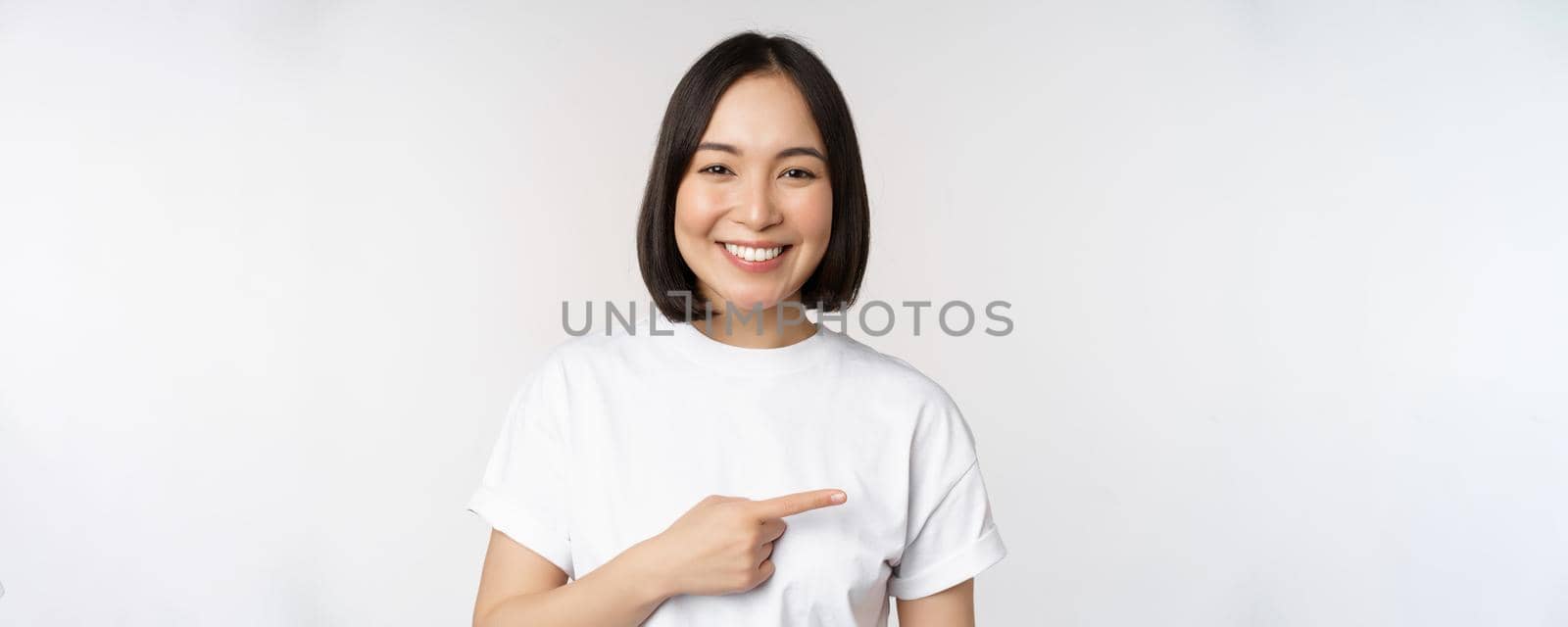 Happy beautiful asian girl showing advertisement on empty copy space, pointing finger right and smiling, demonstrating banner or company logo, white background by Benzoix