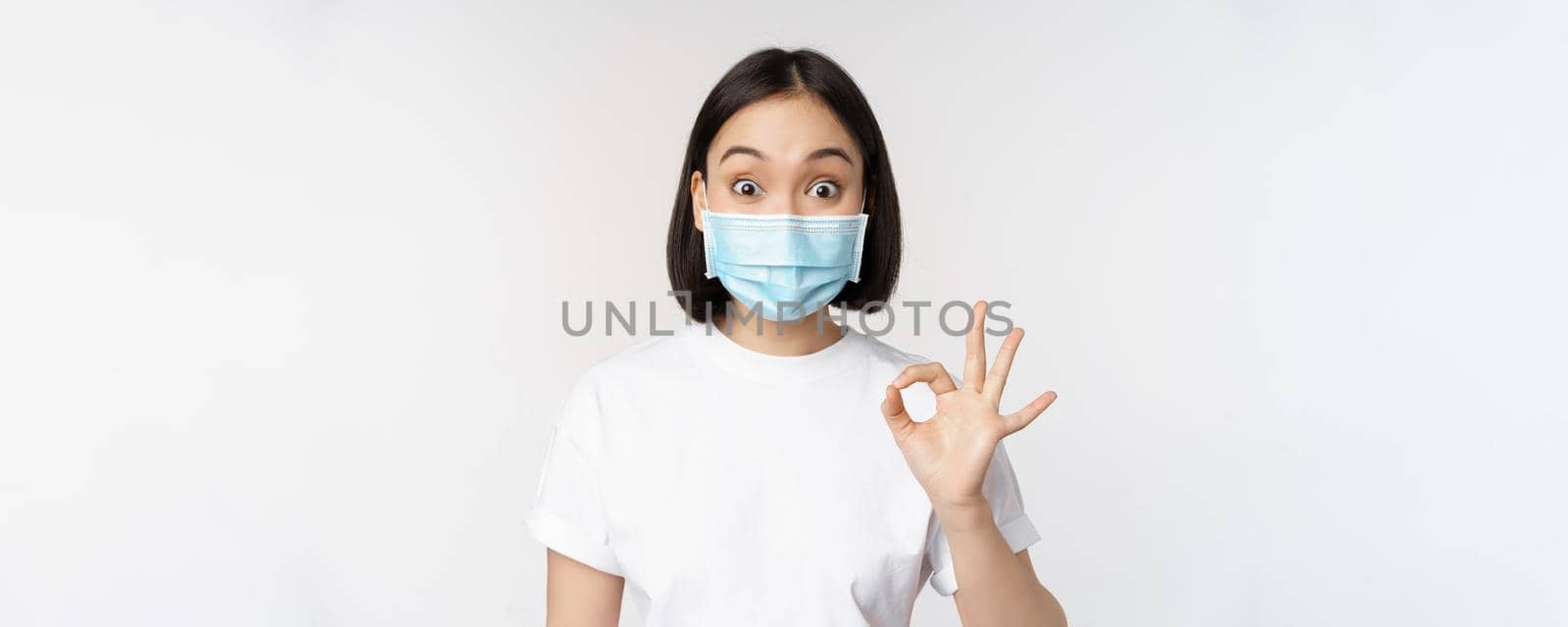 Covid-19, healthcare and medical concept. Impressed asian woman in medical mask, looking amazed and showing ok, okay sign, standing over white background.