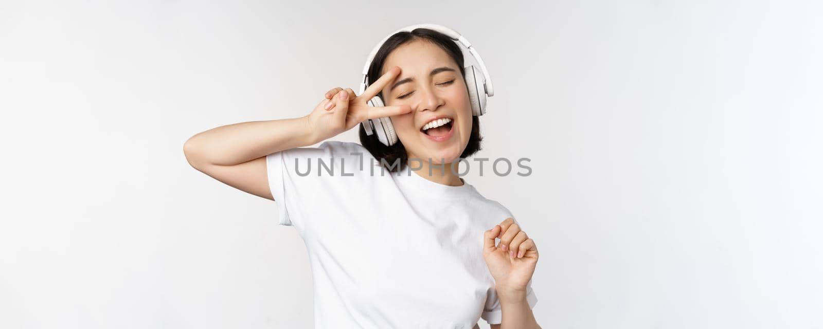 Dancing and singing asian woman, listening music in headphones, standing in earphones against white background by Benzoix