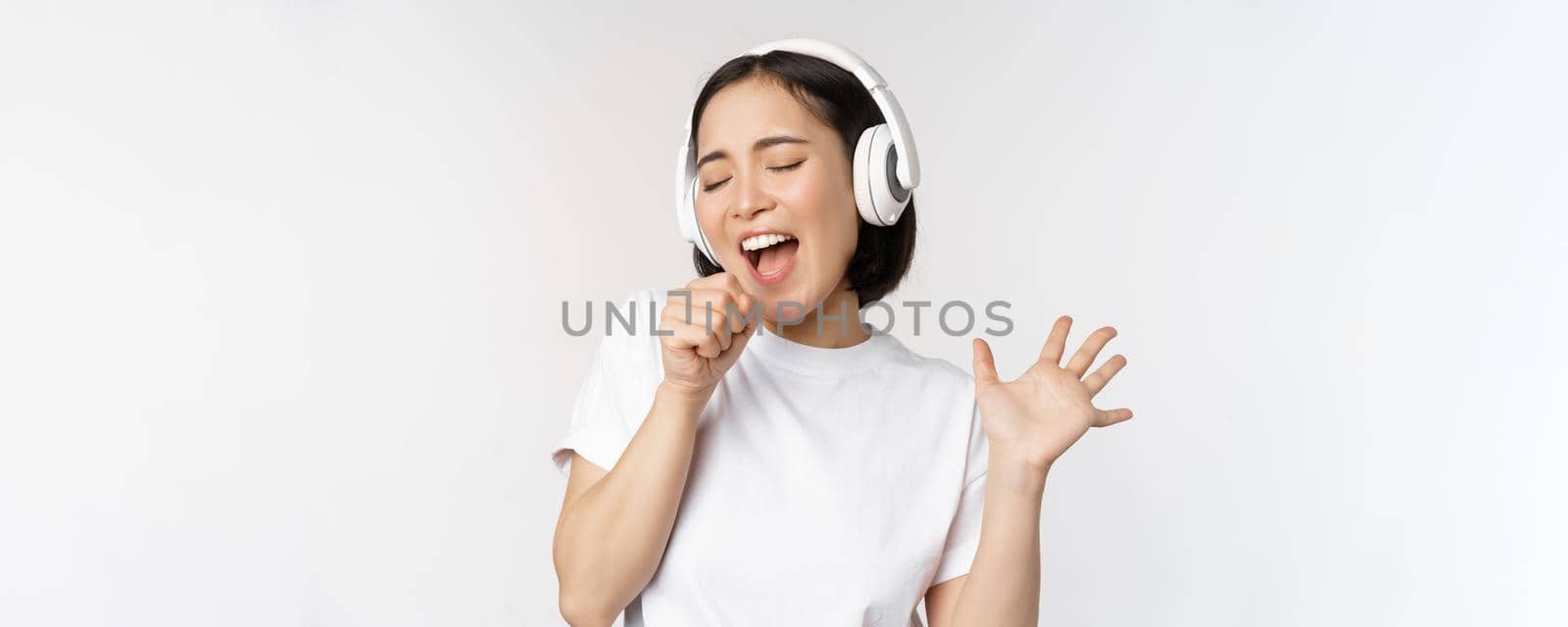 Korean girl sings and listents music in headphones, having fun, stands over white background. Copy space