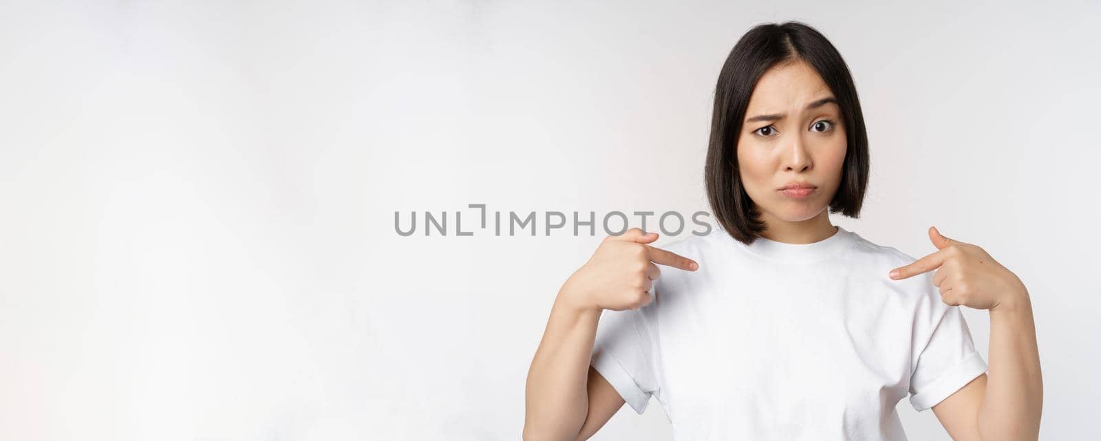 Young asian woman pointing at herself with disbelief, being chosen, surprised by her candidature, standing over white background.