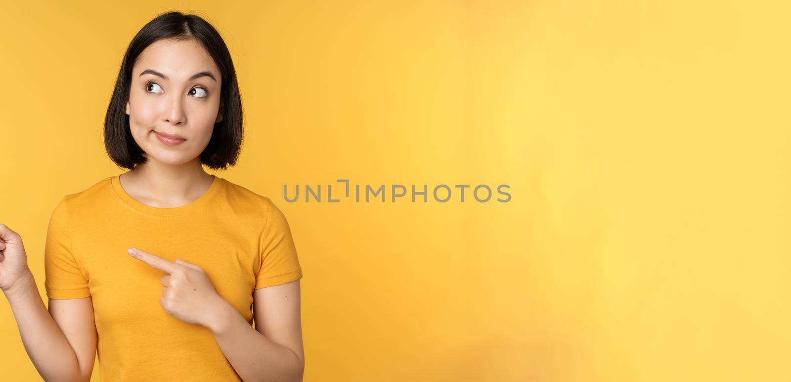 Portrait of intrigued asian woman, looking and pointing fingers left at advertisement, showing smth interesting, standing over yellow background.