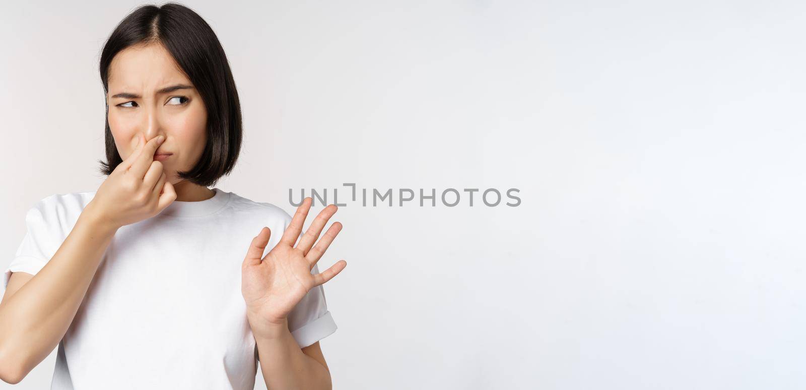 Asian girl looks disgusted, rejecting product with bad smell, shut nose from aversion and cringe, standing against white background.