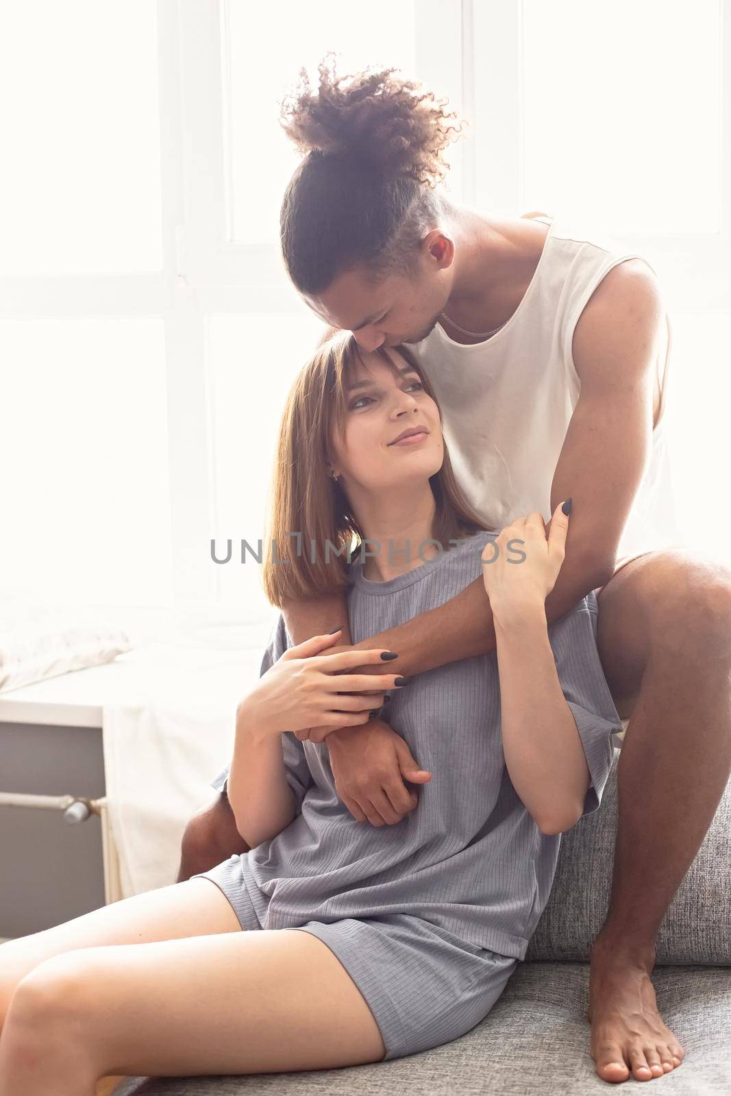 A multiracial couple, a guy and a girl, in cozy home clothes are sitting on a sofa by the window by Zakharova