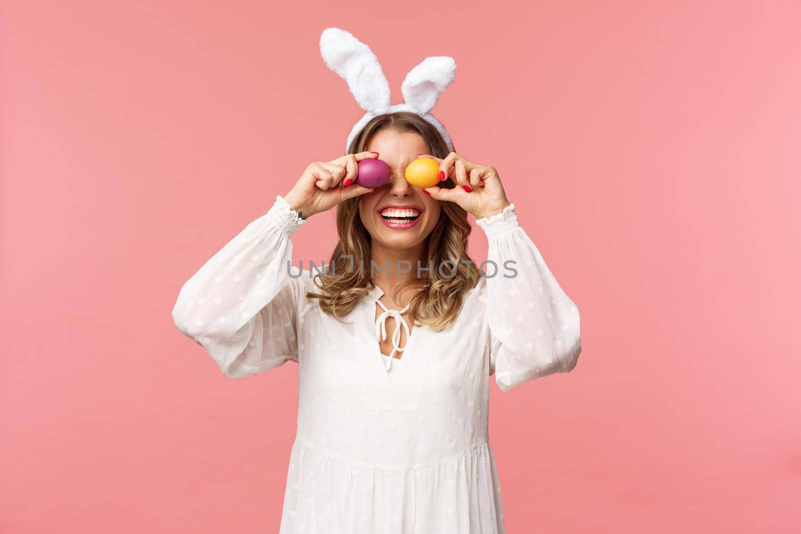 Holidays, spring and party concept. Portrait of lovely, tender smiling woman in rabbit ears and white dress celebrating Easter day, holding painted eggs on eyes and grinning, pink background.