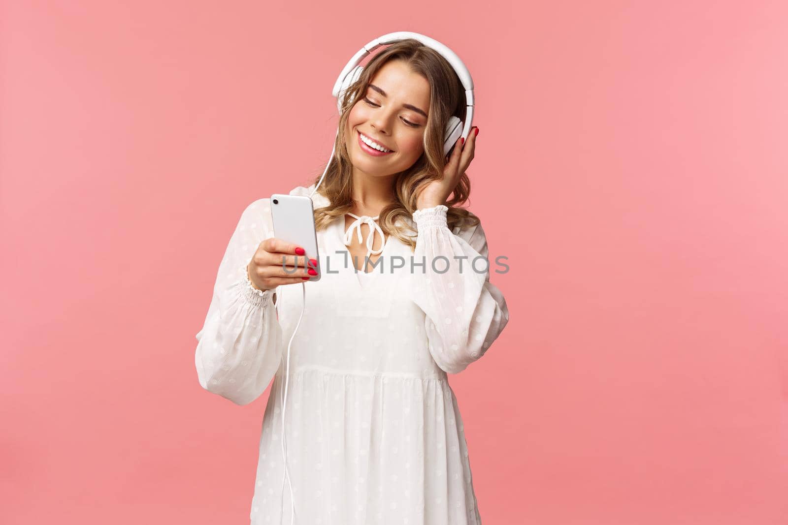 Portrait of dreamy cute and tender blond girl in white dress, holding smartphone, listen music in headphones, smiling at display mobile phone as picking playlist for spring mood, pink background.