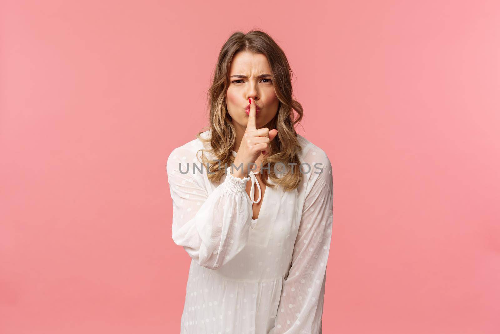 Beauty, fashion and women concept. Angry serious-looking blond girl in white dress, shushing at someone making noise, press finger to lips scolding bad behaviour, say be silent, pink background.