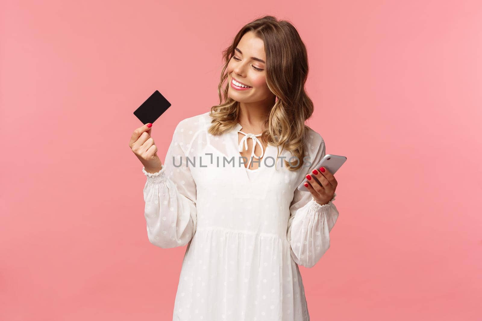 Portrait of happy good-looking young blond woman in white dress, looking pleased and satisfied at credit card, holding smartphone, recommend buy online, internet shopping concept.