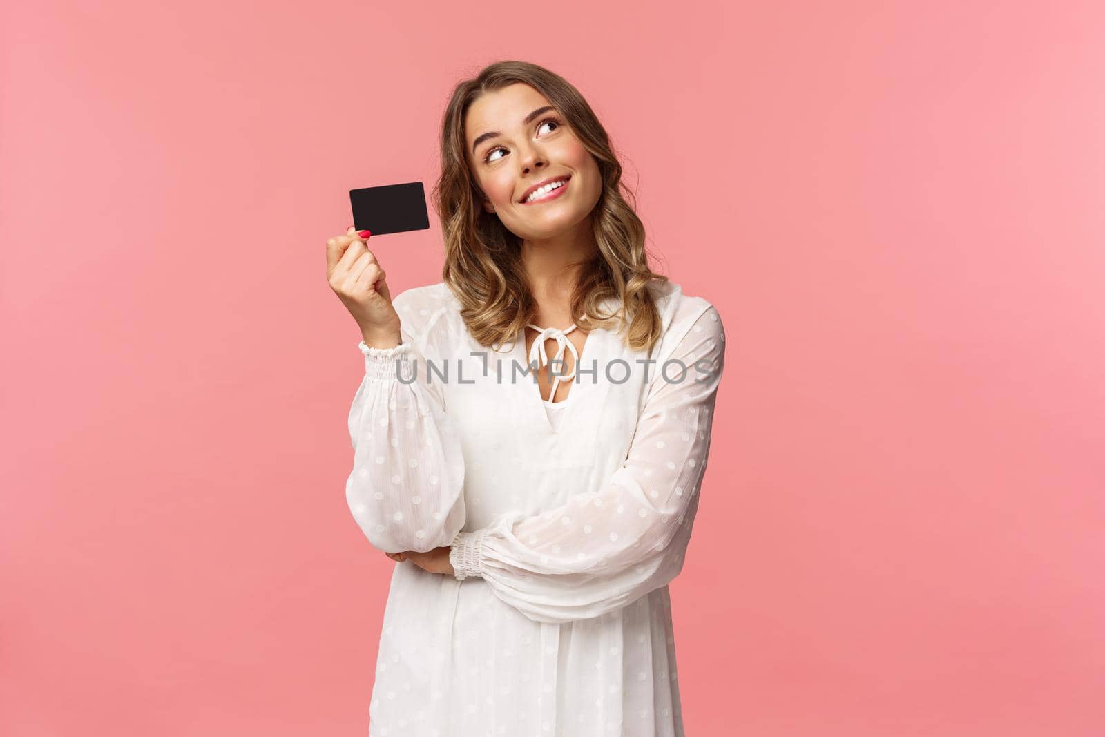 Portrait of cute tender feminine blond girl daydreaming about how she will waste her money on online shopping, look up dreamy, imaging as waiting delivery, hold credit card, pink background.