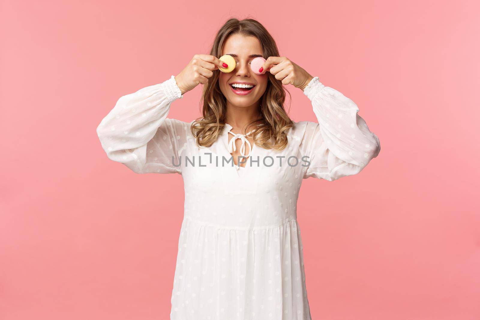 Holidays, spring and party concept. Portrait of carefree funny and cute blond girl in white dress, holding two macarons on eyes and smiling happy, like eating tasty desserts, pink background by Benzoix