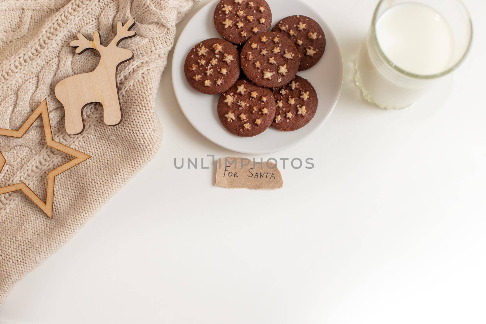 on the table is a saucer with round chocolate chip cookies with stars, a glass of milk, a knitted beige blanket and as well as a small piece of paper with the words -For Santa.