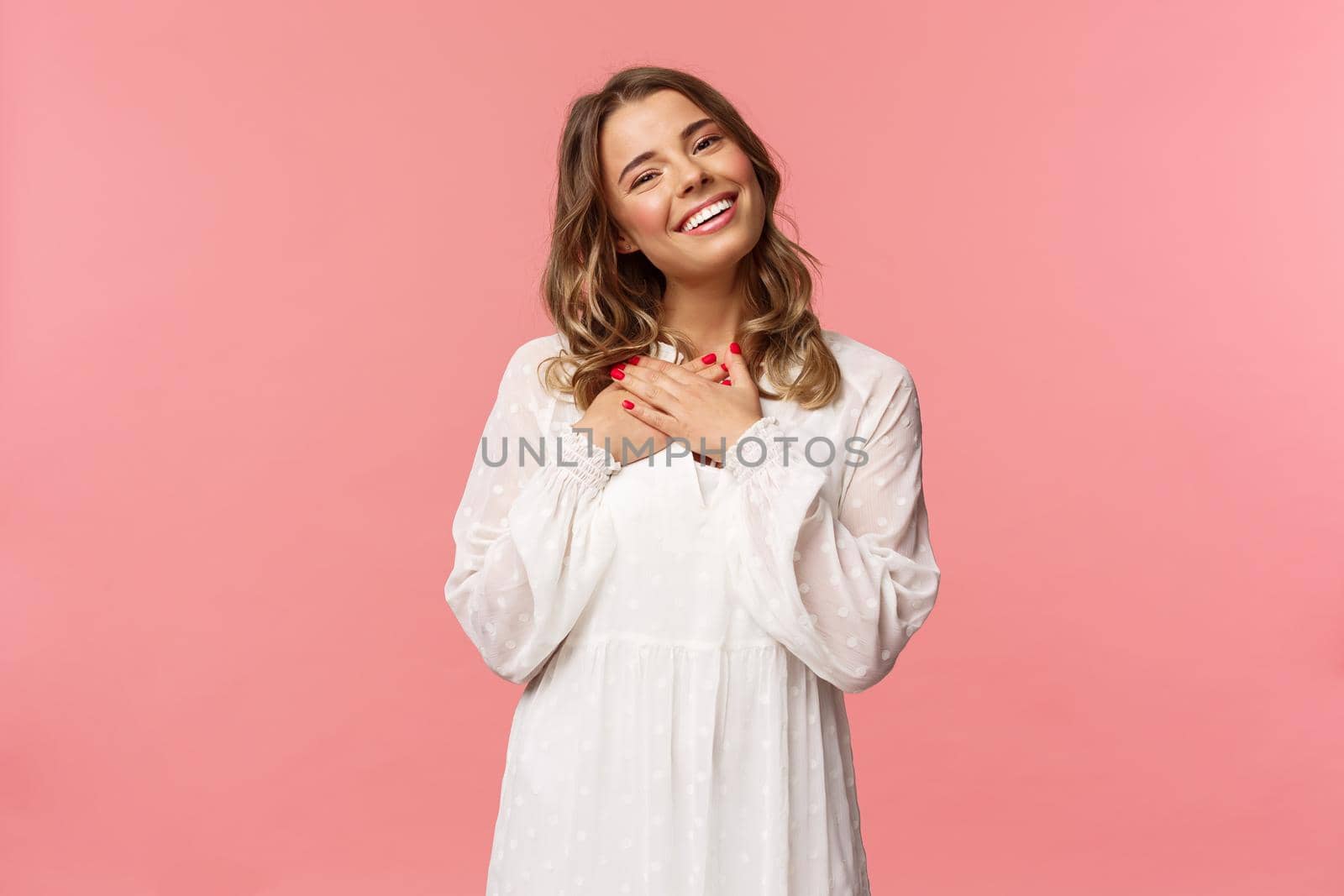 Beauty, fashion and women concept. Portrait of feminine blond young woman in white cute dress, hold arms pressed to heart and sighing from admiration and delight, feel thankful, pink background by Benzoix