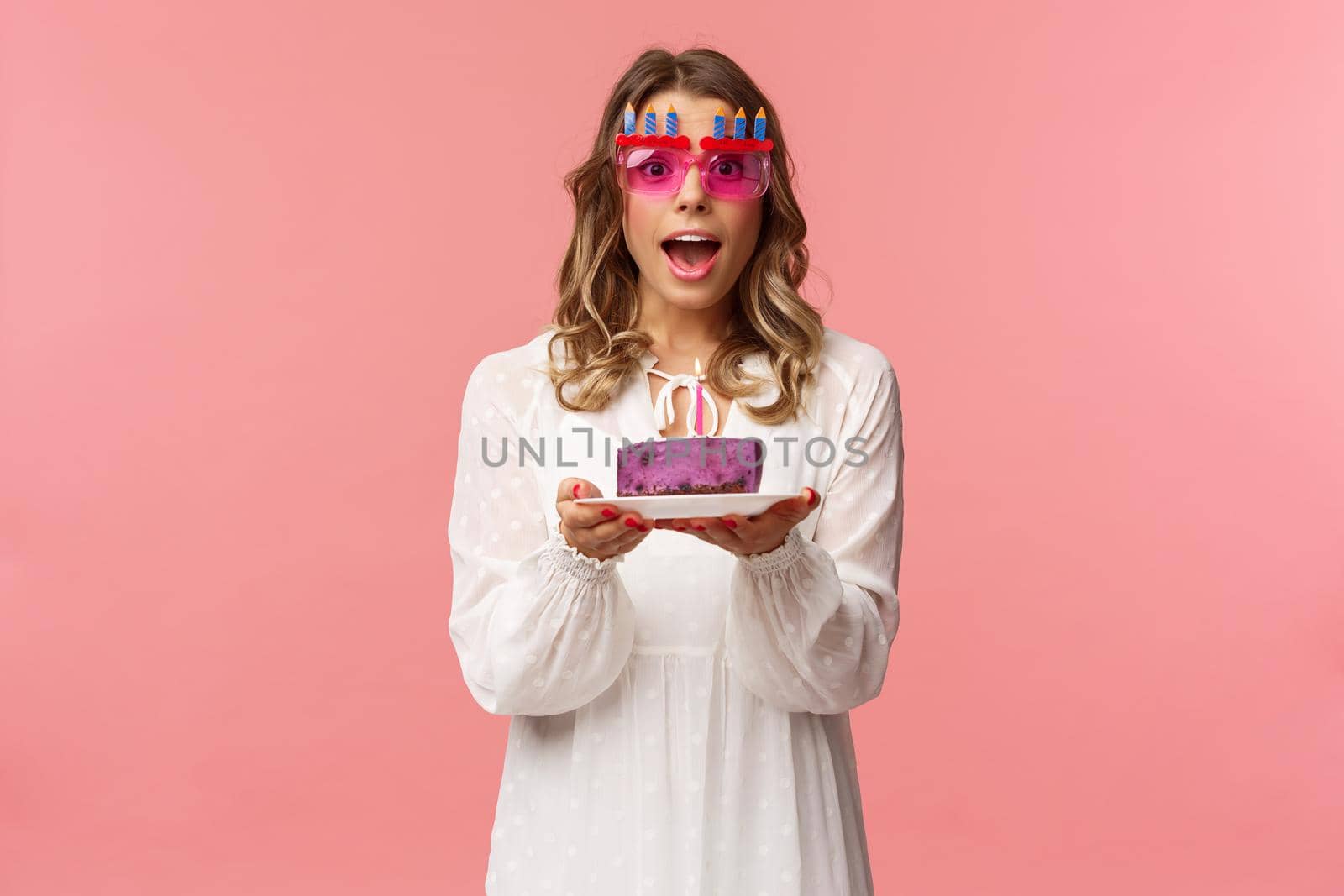 Holidays, spring and party concept. Excited, happy attractive blond woman in white dress, wearing funny b-day glasses, holding birthday cake with lit candle, making wish smiling camera.