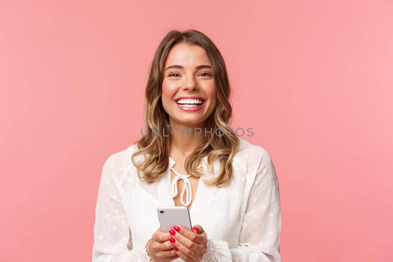Close-up portrait of charming, lovely blond girl using mobile phone, texting friend, order in online store using smartphone application, laughing and smiling pleased, stand pink background by Benzoix