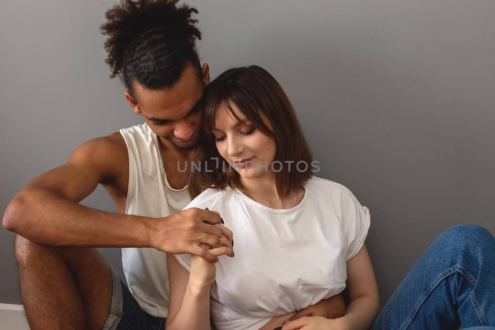 Multiracial couple, guy and girl in white t-shirts sit at home on the floor against a gray wall, hugging, holding hands
