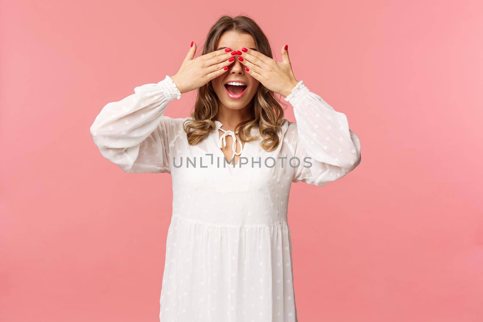 Excited cute blond girl in white spring dress waiting for gifts at surprise party, cover eyes blindfolded standing pink background, open mouth in amazement and anticipation by Benzoix