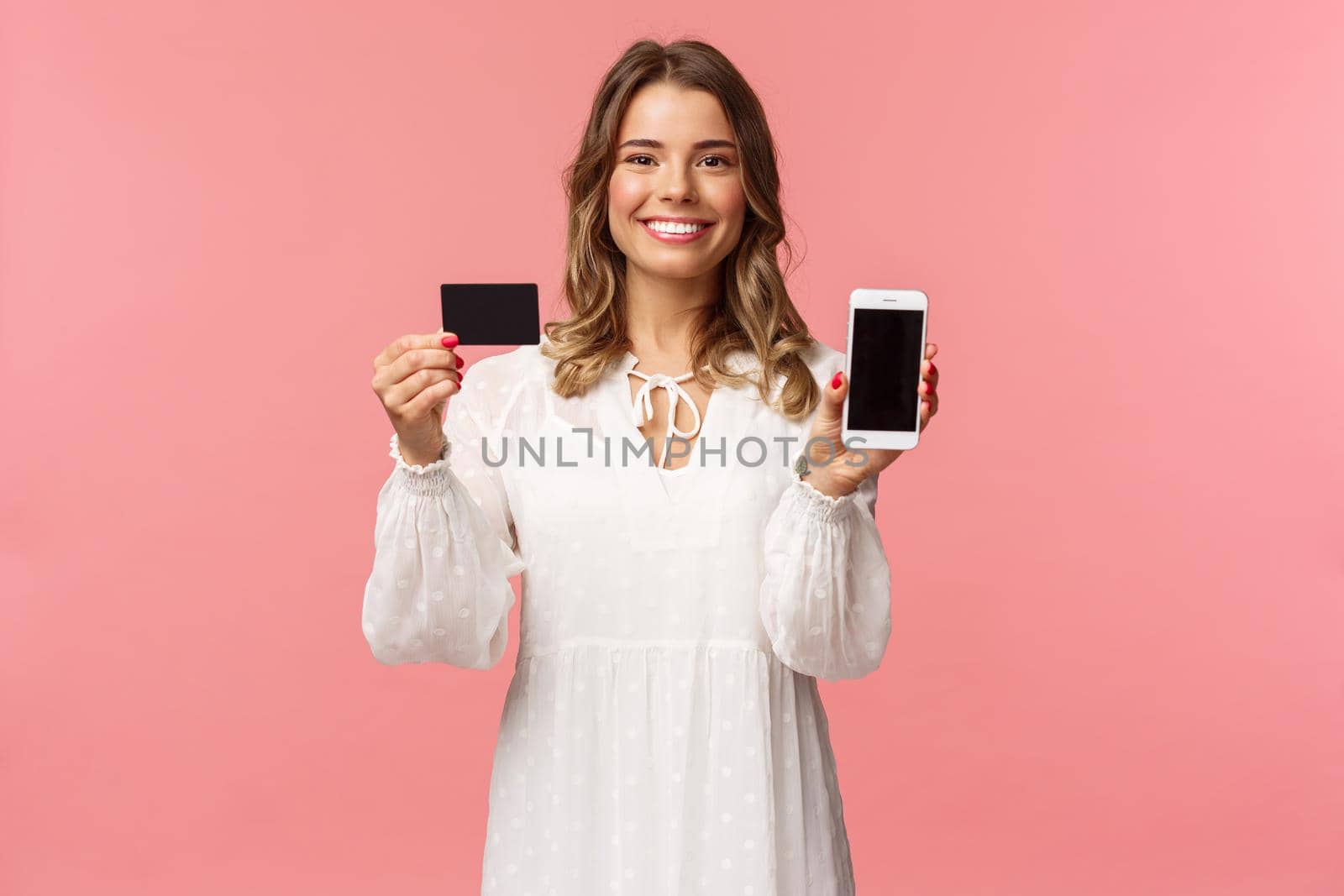 Portrait of satisfied cute blond girl in white dress, showing you her credit card and mobile phone application, online shopping app or bank website, smiling camera, standing pink background.