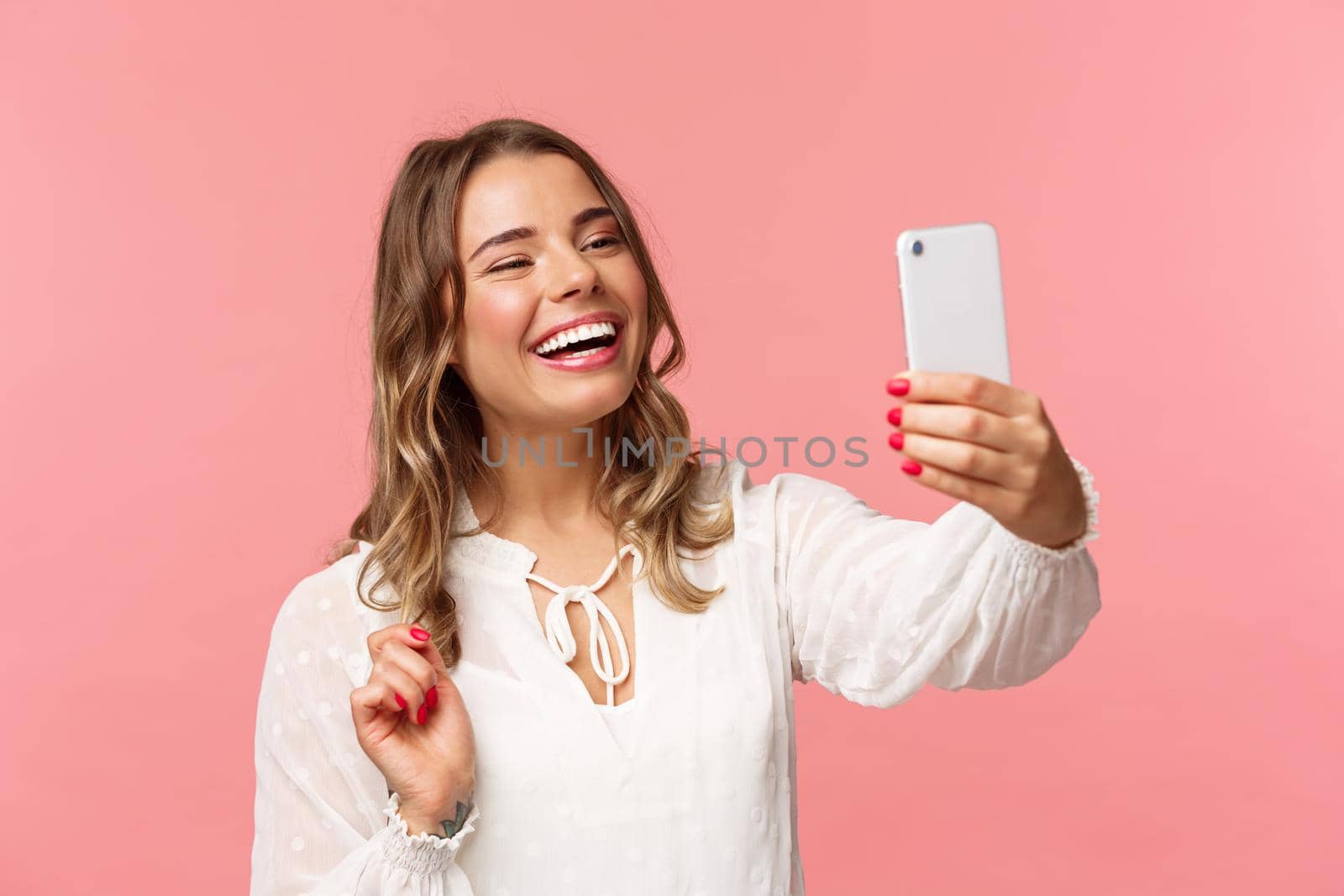 Close-up portrait of cheerful upbeat smiling blond girl, wearing white dress, laughing as record video, calling friend on mobile application, taking photo, selfie on smartphone, pink background.