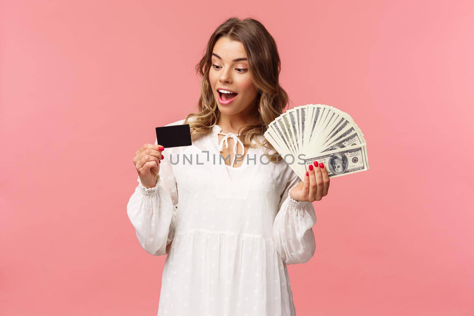 Portrait of rich good-looking blond girl in white dress, holding dollars money and credit card, winning lottery, think how invest, standing happy pink background. Copy space