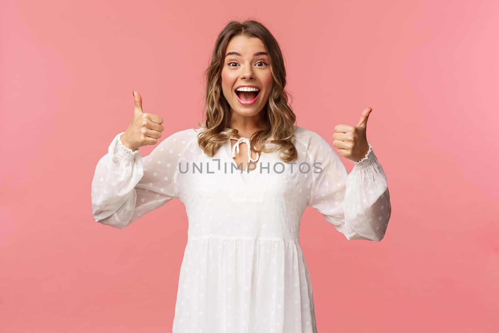 Portrait of excited feminine blond girl in white dress, show thumbs-up in approval, like being pleased, nod agreement, smiling fascinated, leave positive review, recommend product, pink background.