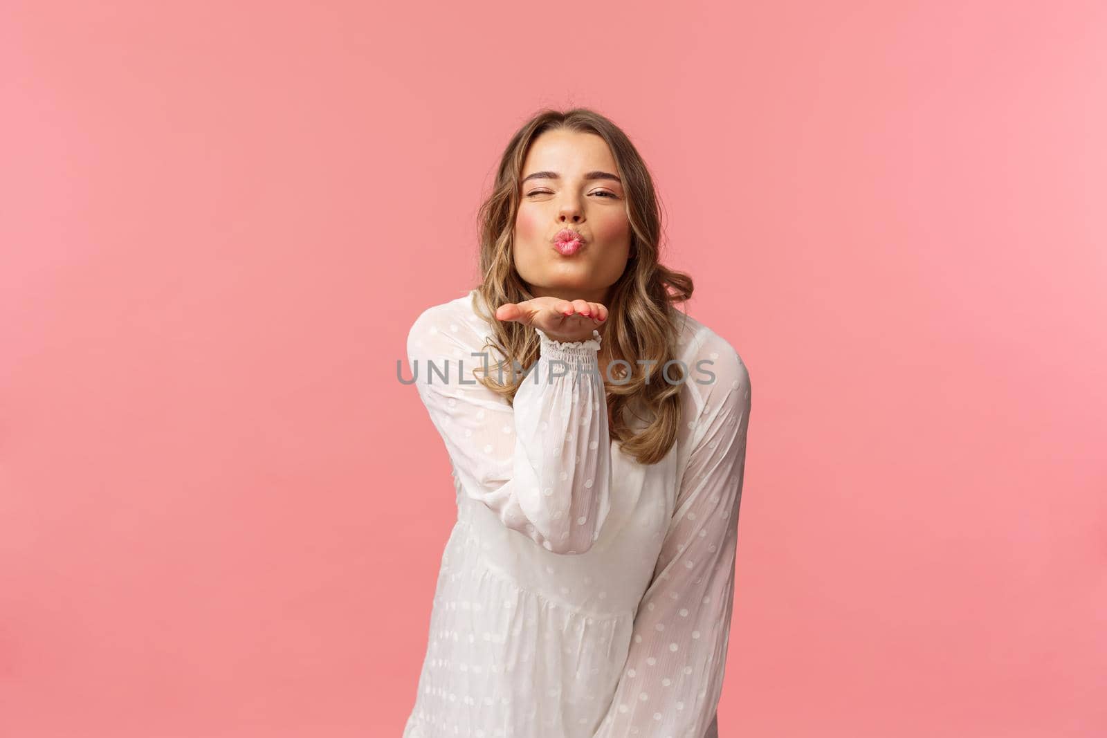 Beauty, fashion and women concept. Portrait of romantic tender, young blond woman in white trendy dress, sending air kiss at camera with sassy flirty smile, hold hand near lips, pink background.