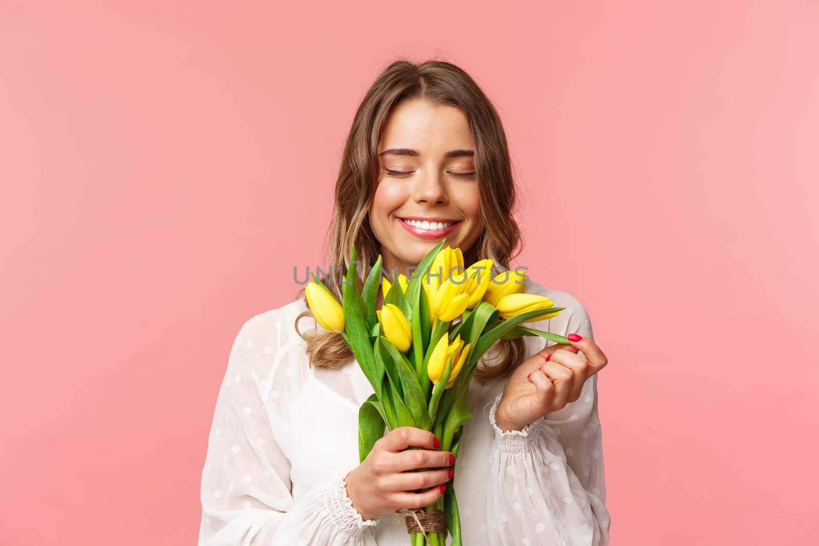 Spring, happiness and celebration concept. Close-up portrait of lovely, romantic blond girl sniffing smell of beautiful yellow tulips, close eyes and smiling happy, standing pink background.