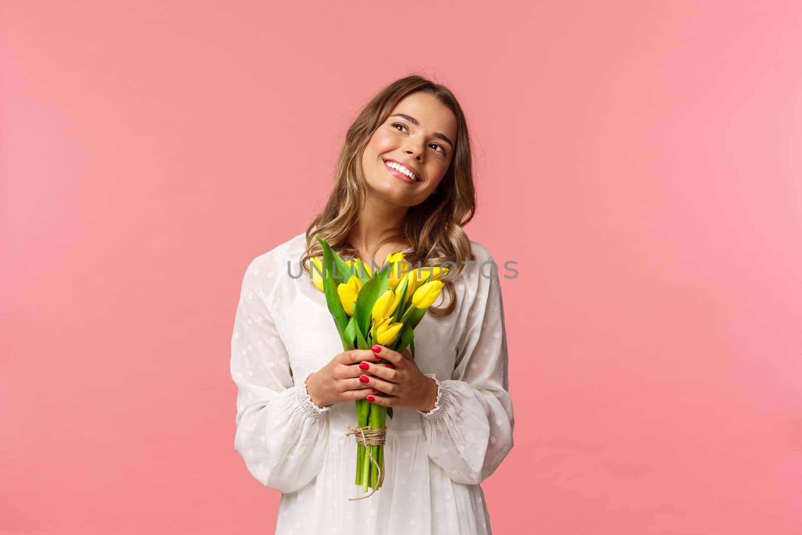 Holidays, beauty and spring concept. Portrait of dreamy, happy blond girl feeling romantic daydreaming about her girlfriend, holding yellow tulips, wear white dress, standing pink background.