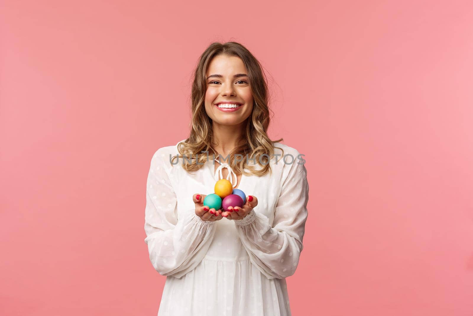 Holidays, spring and party concept. Portrait of tender, lovely blonde girl in white dress, holding Easter painted eggs, celebrating orthodox day, smiling cheerful share positivity, pink background.