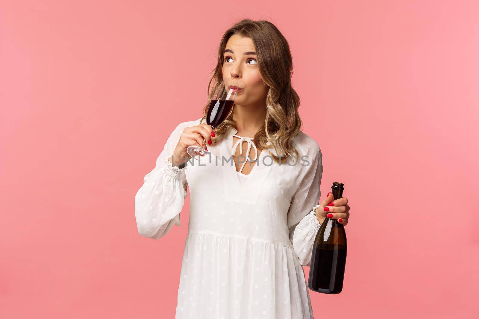Holidays, spring and party concept. Portrait of carefree independent cute blond woman sipping wine from glass, holding bottle and tasting drink, look up, celebrating with friends, pink background.
