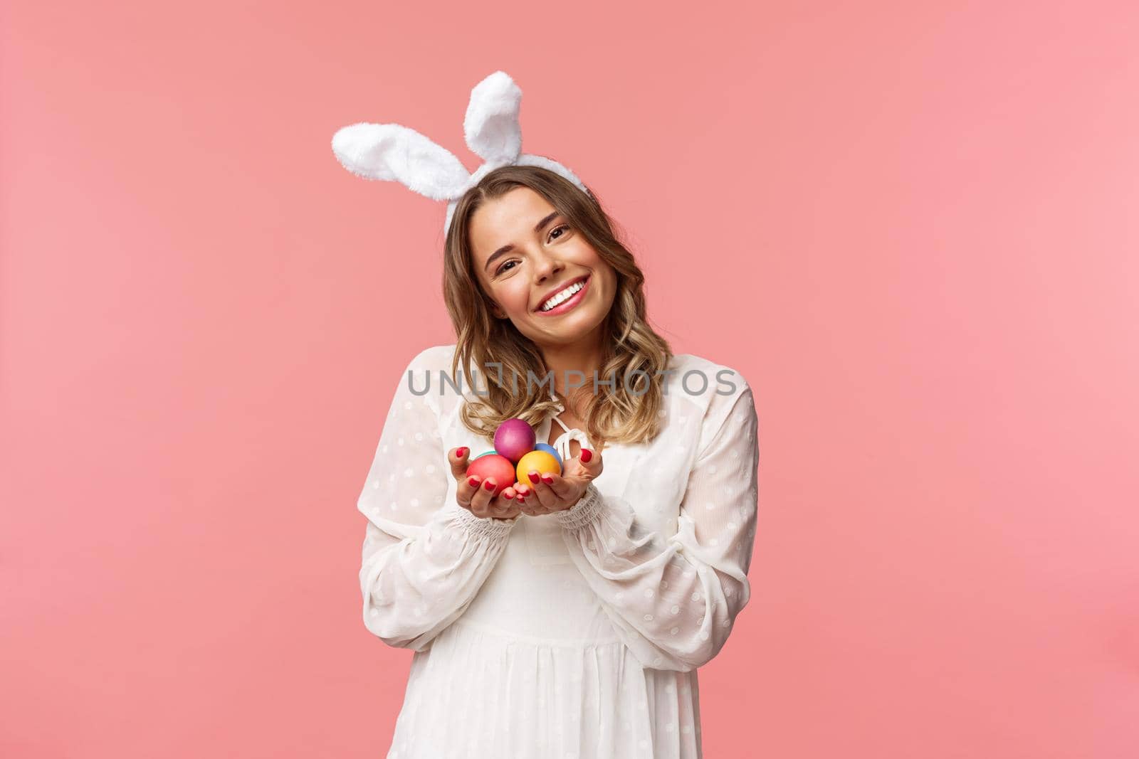 Holidays, spring and party concept. Portrait tender, romantic blond girl in white dress and rabbit ears, tilt head cute, smiling happy as holding painted eggs, celebrating Easter, pink background.