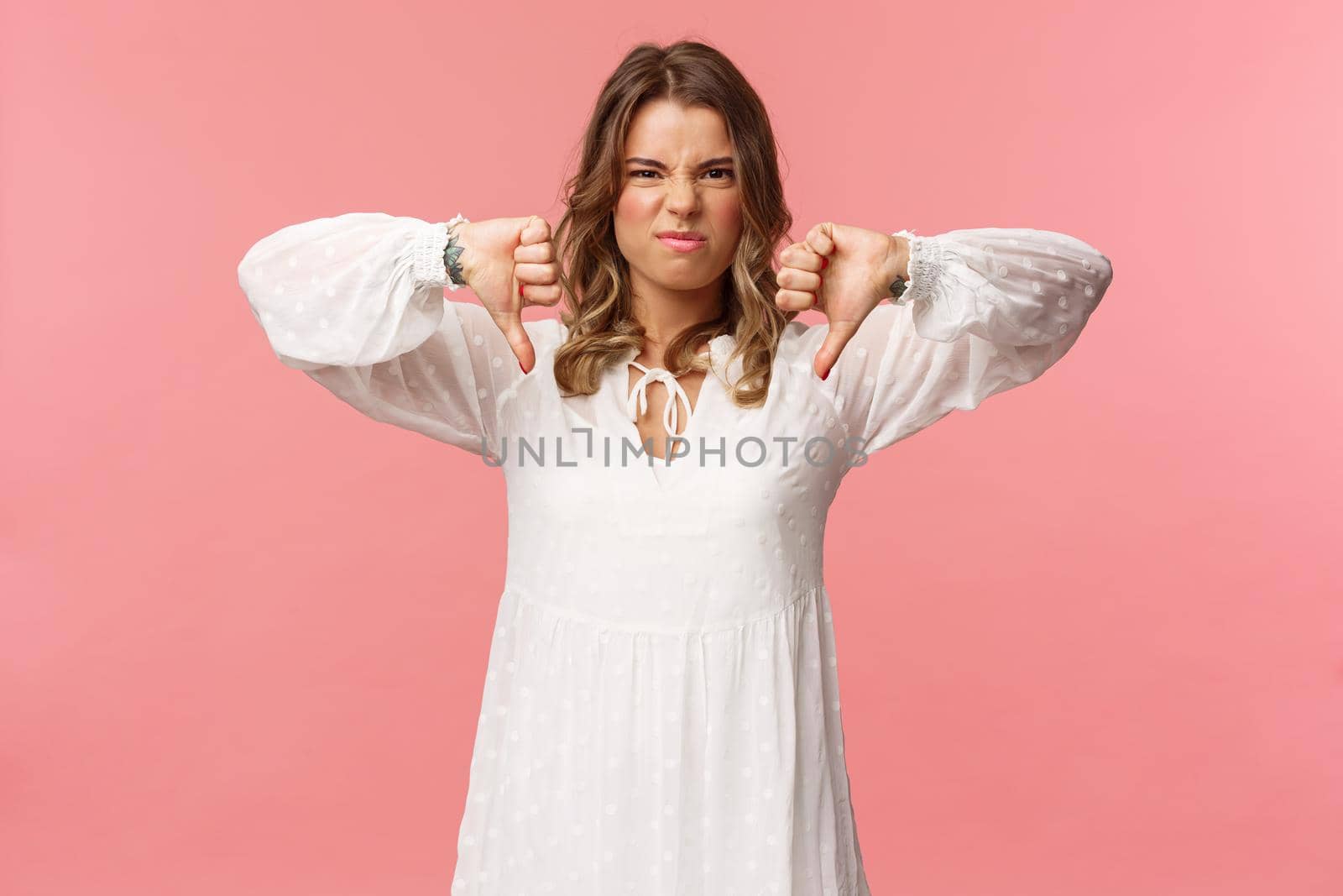 Portrait of picky young blond caucasian woman in white dress over pink background expressing dislike, show thumbs-down and grimacing in aversion, disappointed with bad quality.