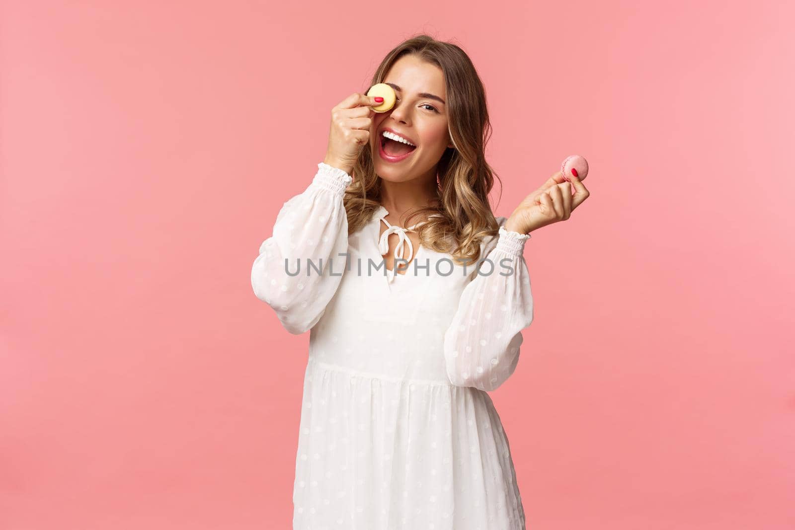 Holidays, spring and party concept. Portrait of carefree, tender and feminine girl in white dress, holding macarons dessert over eye, feeling cheerful, eating sweets, tasty desserts, pink background.