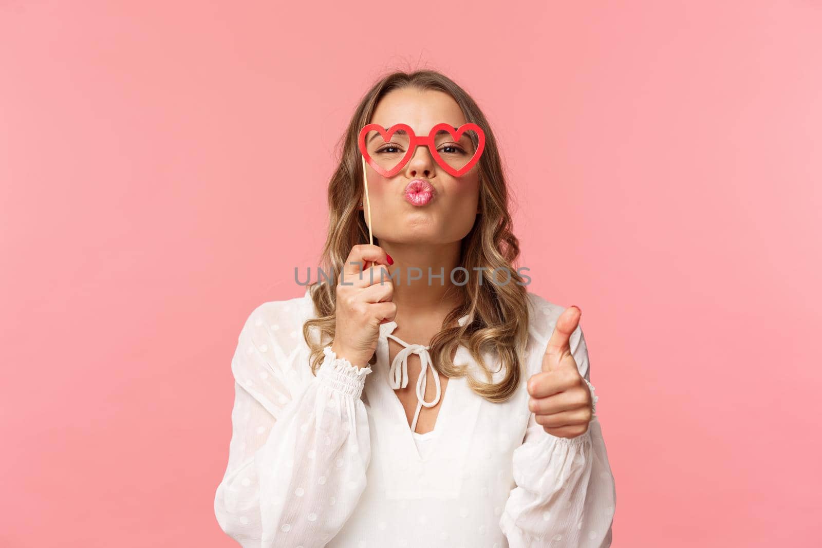 Spring, happiness and celebration concept. Close-up portrait sassy and coquettish attractive blond girl in white dress, holding heart-shaped glasses on eyes, folding lips in kiss and show thumbs-up.