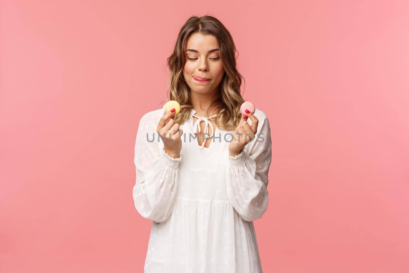 Holidays, spring and party concept. Portrait of cute romantic blond girl in white dress, licking lips as tempting to eat delicious dessert, holding two macarons and look pleased, pink background by Benzoix