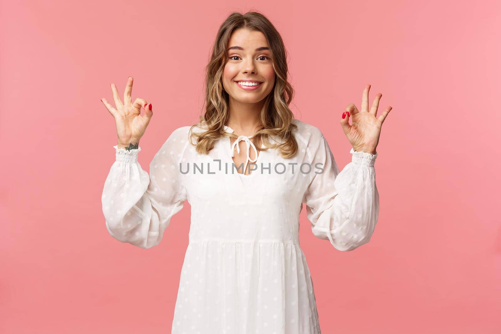 Beauty, fashion and women concept. Portrait of excited and upbeat blond female in white dress, standing satisfied over pink background, show okay sign, guarantee and assure in quality.