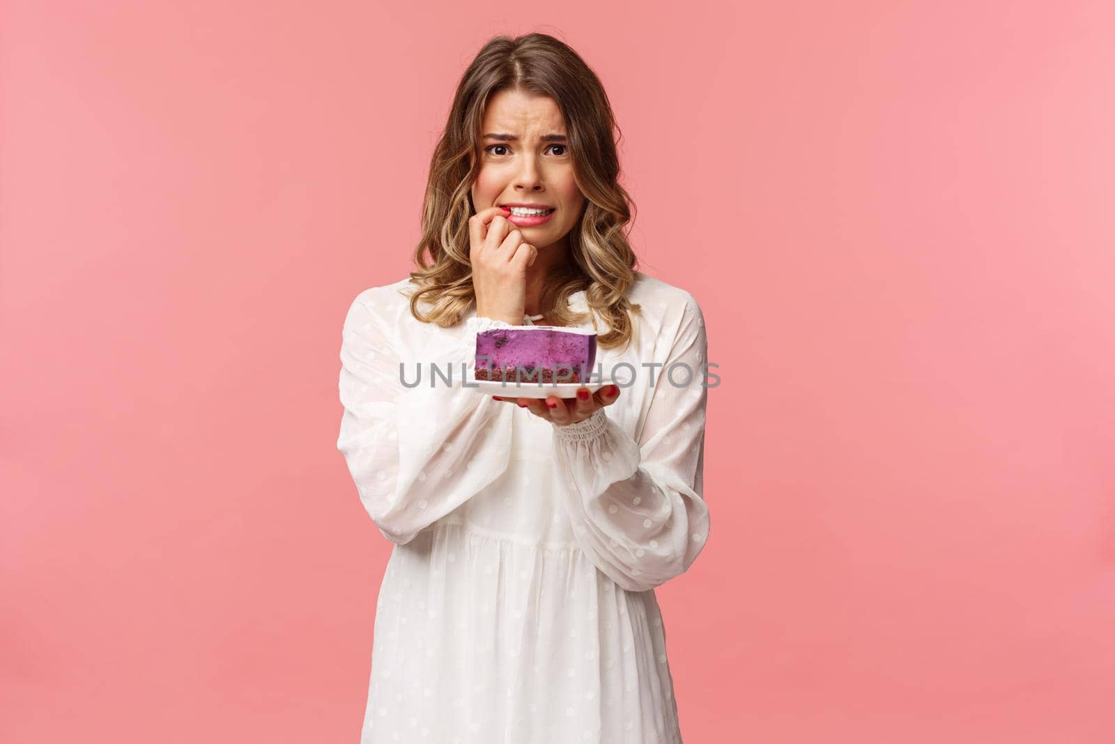 Holidays, spring and party concept. Alarmed, worried young blond girl hesitating eat cake or not, being on diet, trying stay healthy, holding dessert, biting finger nervously, pink background.